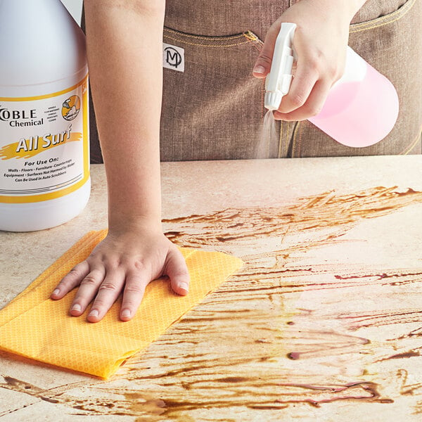 A person cleaning a counter with a spray bottle of Noble Chemical All Surf All Purpose Cleaner.