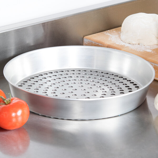 An American Metalcraft silver tin-plated pizza pan with holes in it sitting next to a ball of dough and tomatoes.