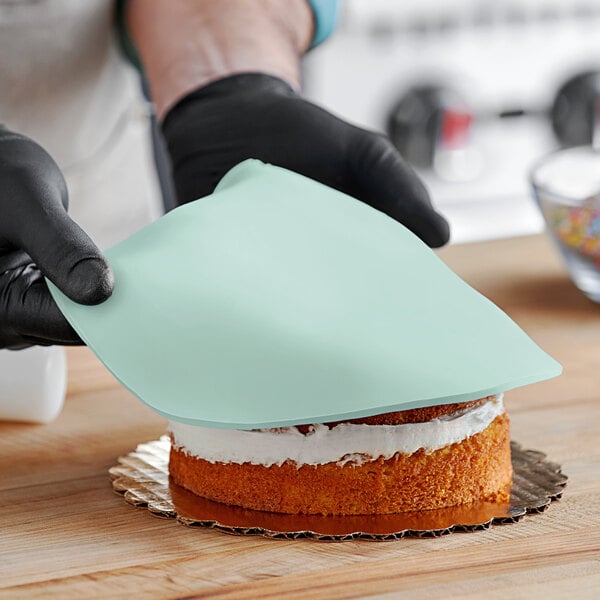 A person wearing black gloves and using a blue cloth to smooth Satin Ice Pastel Green Vanilla Rolled Fondant over a cake.