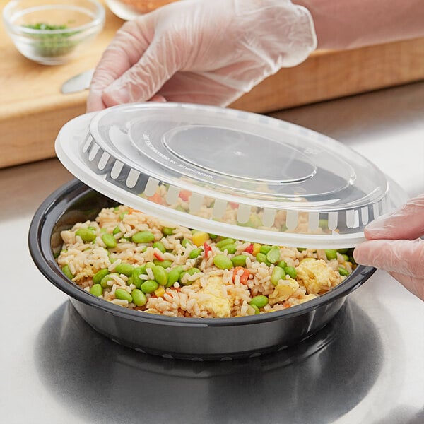 A gloved hand places a black plastic lid on a bowl of rice and vegetables.