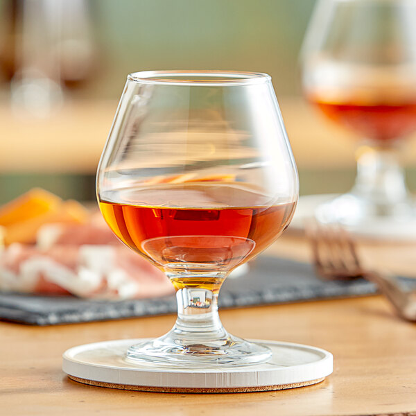 An Acopa Select brandy snifter filled with brown liquid on a table with a coaster.