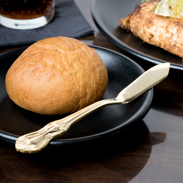 A bread roll and a 10 Strawberry Street Crown Royal gold plated stainless steel butter knife on a black plate.