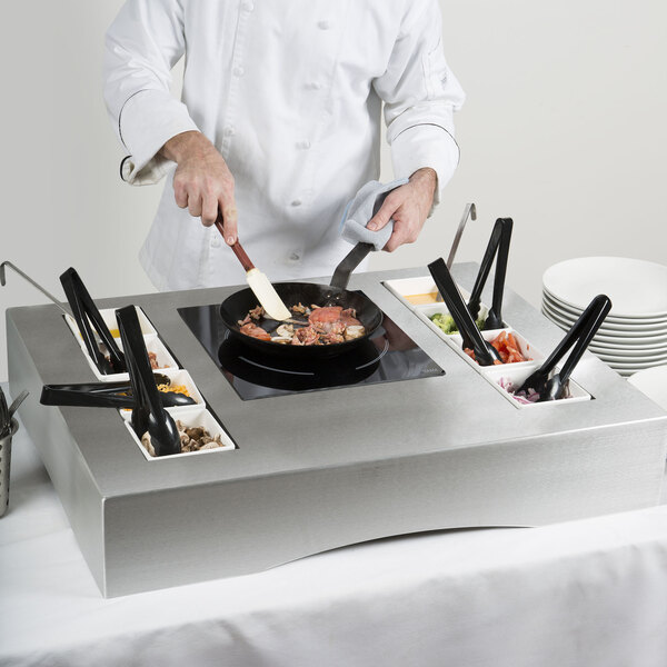 A chef cooking food in a Tablecraft brushed aluminum induction station kit on a countertop.