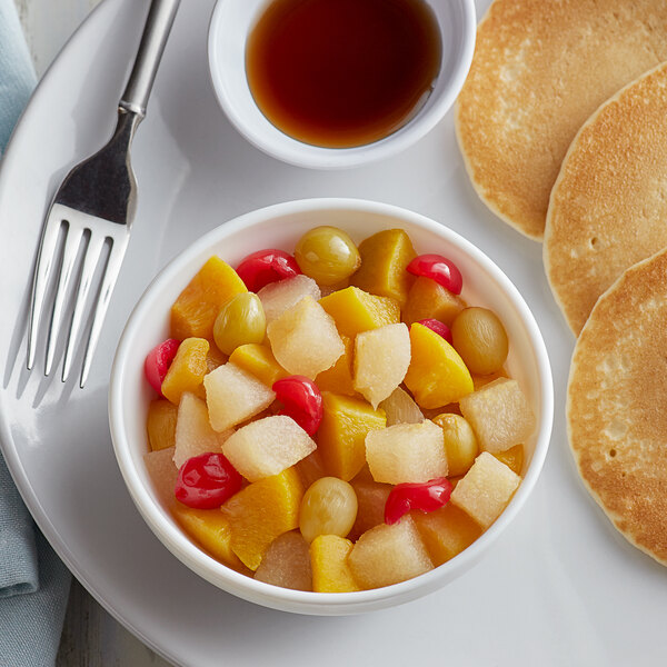 A bowl of fruit salad with yellow fruit and syrup on a table with a cup of brown liquid and a plate of pancakes.
