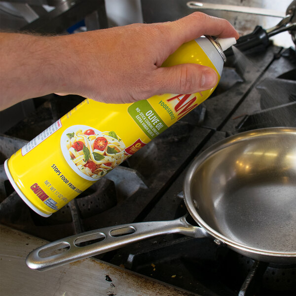 A hand using PAM Olive Oil Release Spray on a pan on a stove.