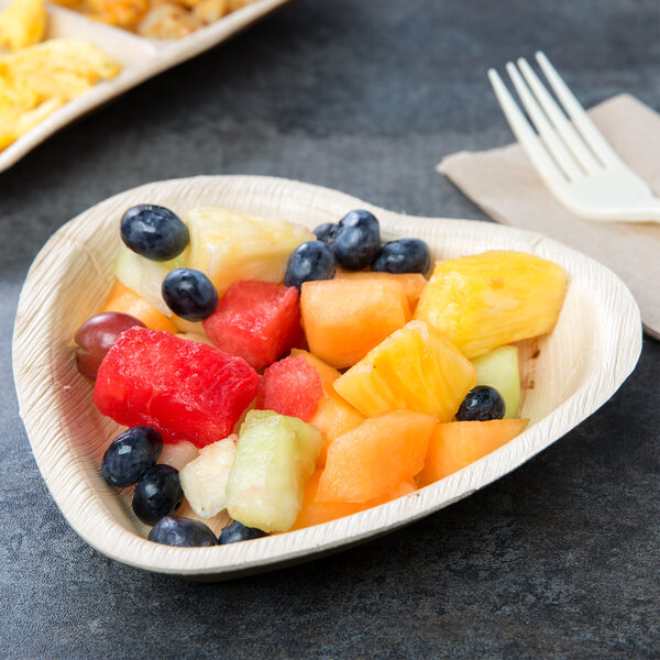 A TreeVive compostable palm leaf heart shaped plate with a fork and knife with a plate of fruit on a table.