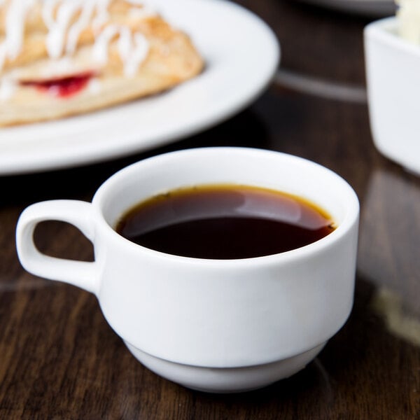 A stackable white porcelain espresso cup filled with coffee on a table with a pastry.