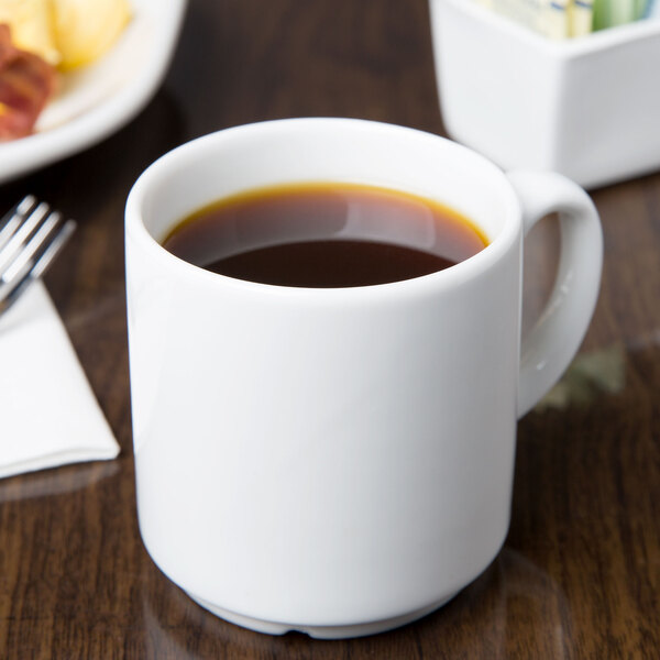 A white Libbey Reflections stackable mug filled with brown liquid on a table with a plate of food.
