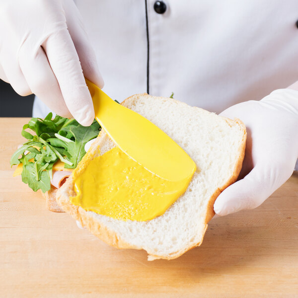 A person in white gloves is using a yellow HS Inc. polypropylene sandwich spreader to spread mustard on a sandwich.
