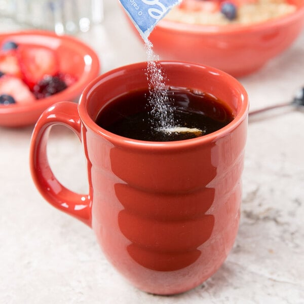 A person pouring sugar from a bag into a red Libbey porcelain mug.