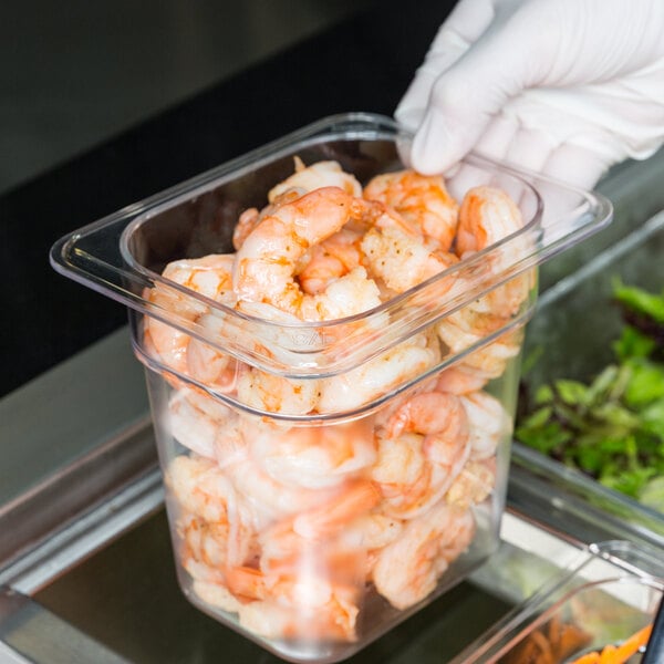 A hand in a glove holding a Cambro clear plastic food pan with shrimp inside.