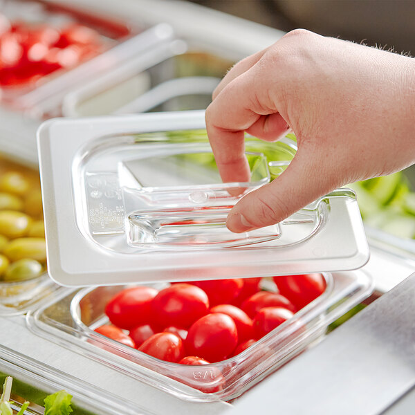 A hand using a Cambro clear plastic lid with a spoon notch to cover a container of tomatoes.