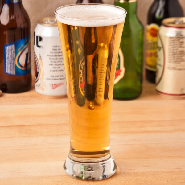 A Libbey pilsner glass filled with beer sitting on a table with other beer bottles.