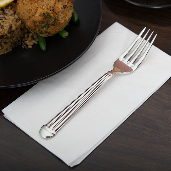 A Libbey stainless steel dinner fork on a napkin next to a plate of food.