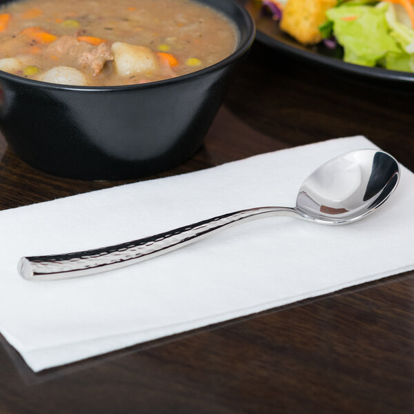 A silver Libbey Chivalry bouillon spoon on a white napkin next to a bowl of soup.