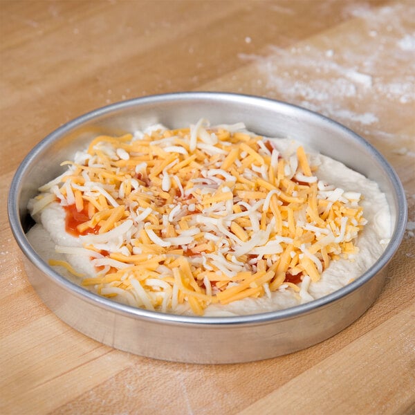 An American Metalcraft aluminum pizza pan with pizza dough, cheese, and sauce in it.