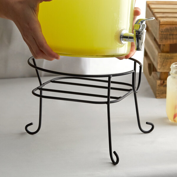 A hand pouring yellow liquid from a yellow container into a black metal stand with a spigot.