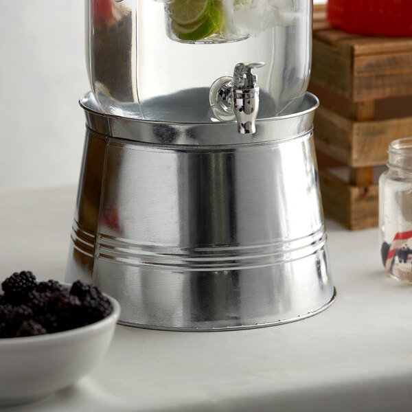 A silver metal base for an Acopa beverage dispenser on a table with a bowl of blackberries.