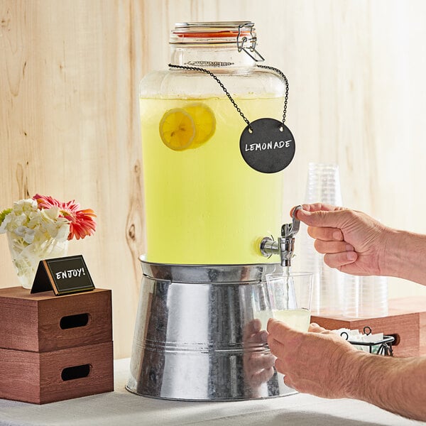 A person pouring lemonade from an Acopa glass dispenser with a jar of lemonade on a table.