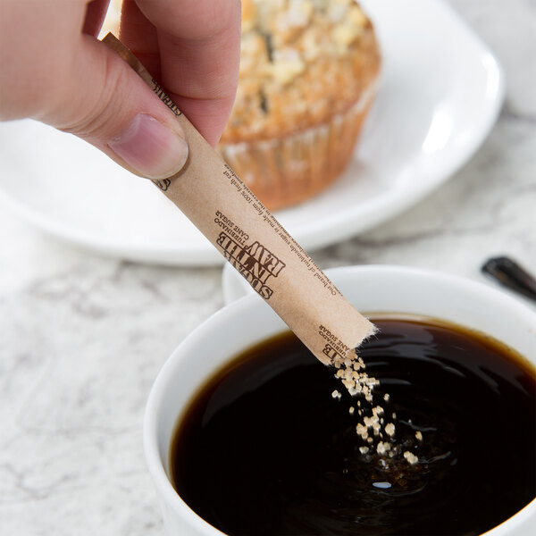 A hand holding a Sugar In The Raw paper tube over a cup of coffee.