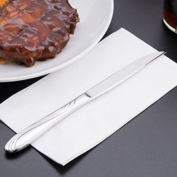 A Libbey Neptune steak knife on a napkin next to a plate of meat.