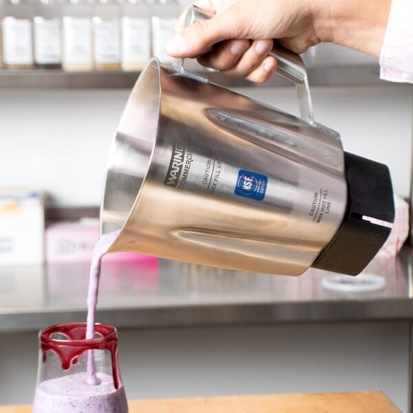 A person pouring a smoothie from a Waring stainless steel blender jar into a glass.