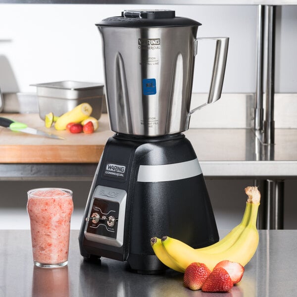 A Waring bar blender with a stainless steel container filled with a pink smoothie, next to a banana and strawberries.