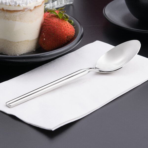 A Libbey stainless steel dessert spoon on a napkin next to a plate of cake.