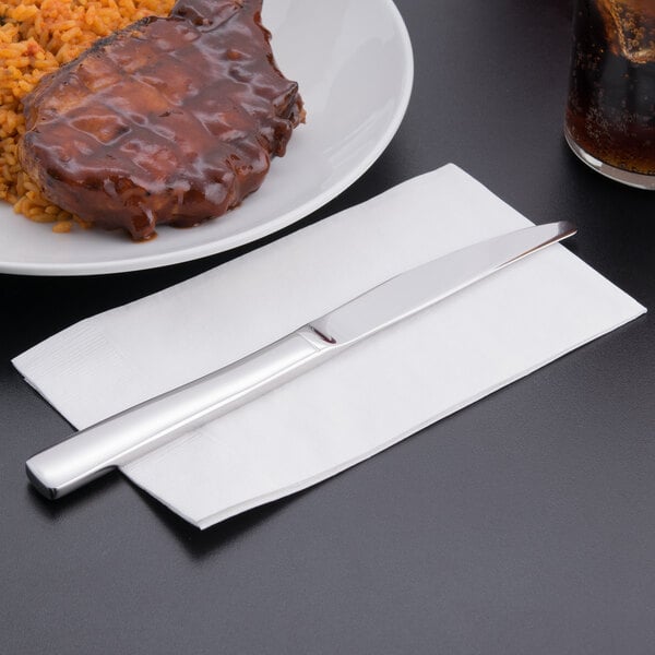 A Libbey stainless steel dinner knife on a napkin next to a plate of meat and rice.
