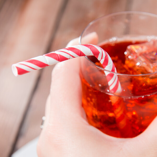 A hand holding a glass of red liquid with a Creative Converting red and white striped paper straw.