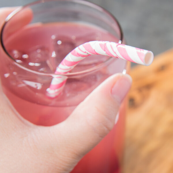 A hand holding a glass of pink liquid with a Creative Converting pink and white striped paper straw.