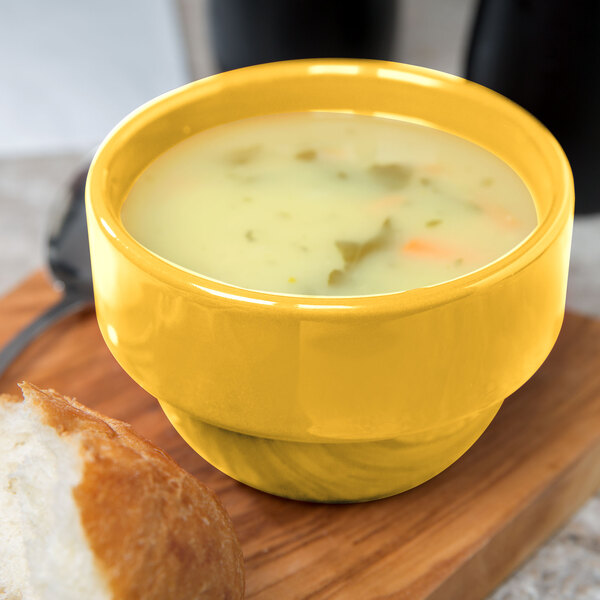 A yellow bowl of Libbey Cantina saffron bouillon soup on a white surface next to a piece of bread.
