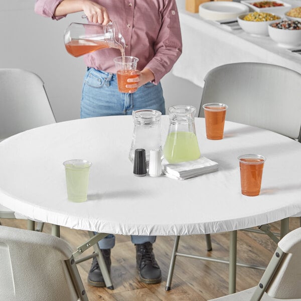 A woman pouring orange liquid from a plastic cup onto a table with a white Stay Put tablecloth.