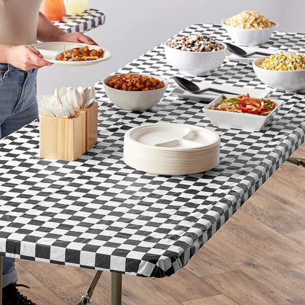 A person standing at a table with a black and white checkered rectangular tablecloth with food on it.