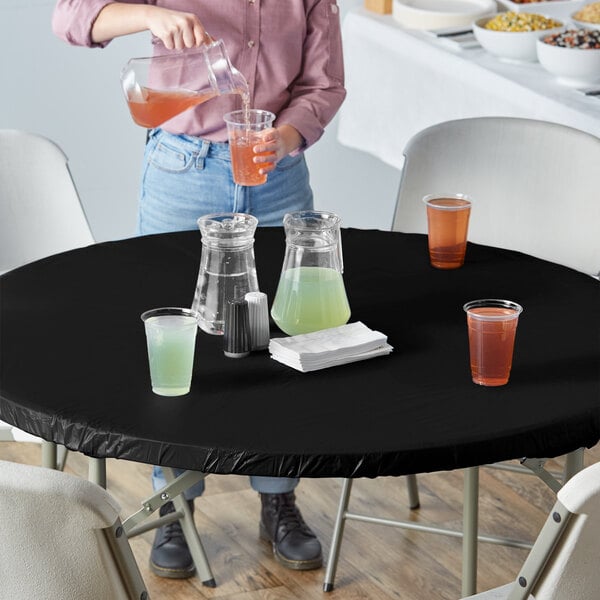 A woman pouring a brown drink into a plastic cup with a straw on a black table with a Creative Converting Stay Put black tablecloth.