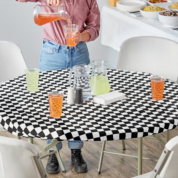 A woman pouring orange liquid into a glass on a black and white checkered table with a bowl of corn.