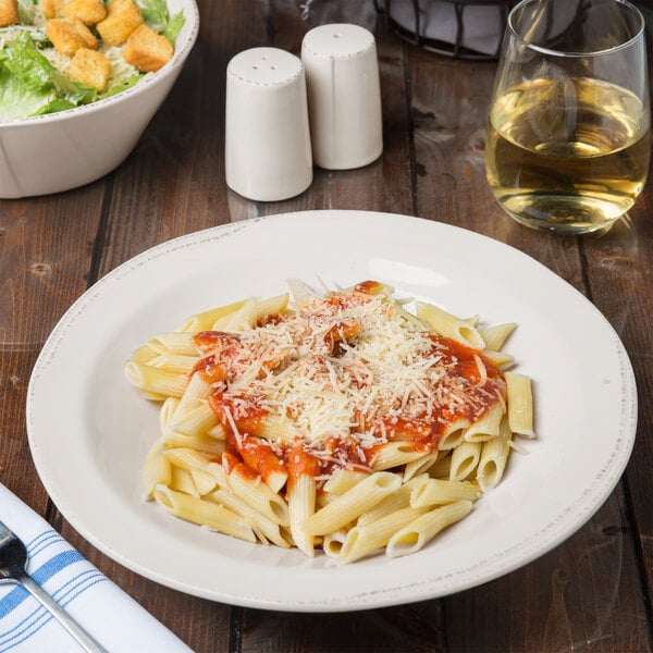 A Libbey ivory porcelain pasta bowl filled with pasta with sauce and a plate of salad with croutons on a table with wine.