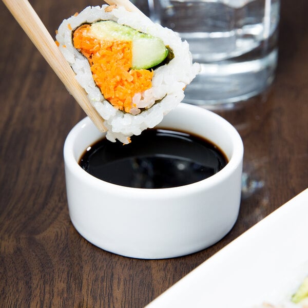 A sushi roll being held by chopsticks and dipped into a Libbey ultra bright white porcelain disk bowl filled with soy sauce.