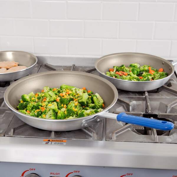 Three Vollrath aluminum non-stick fry pans with blue Cool handles on a stove with vegetables in them.