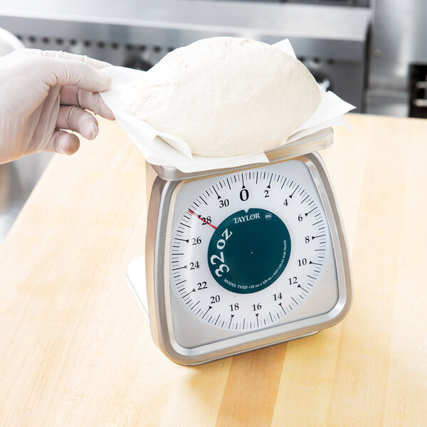 A hand using a Taylor portion scale to weigh a white ball of dough.
