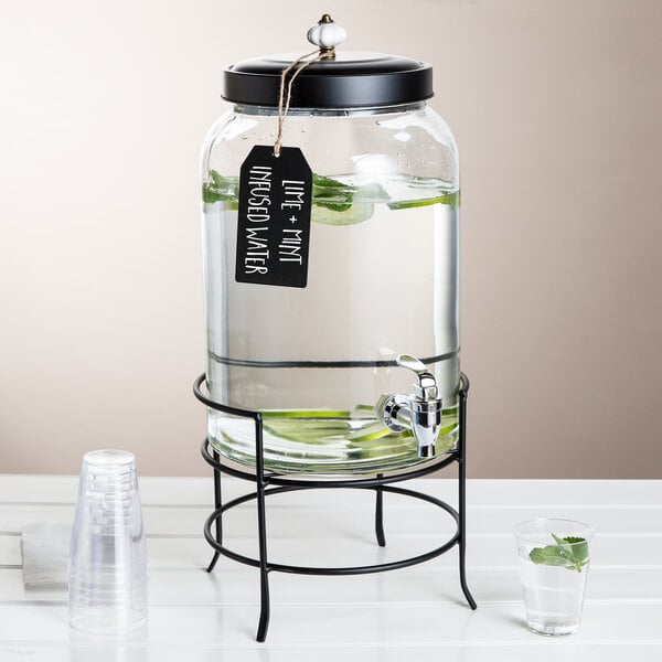 A Stylesetter glass beverage dispenser on a metal stand with water in it.