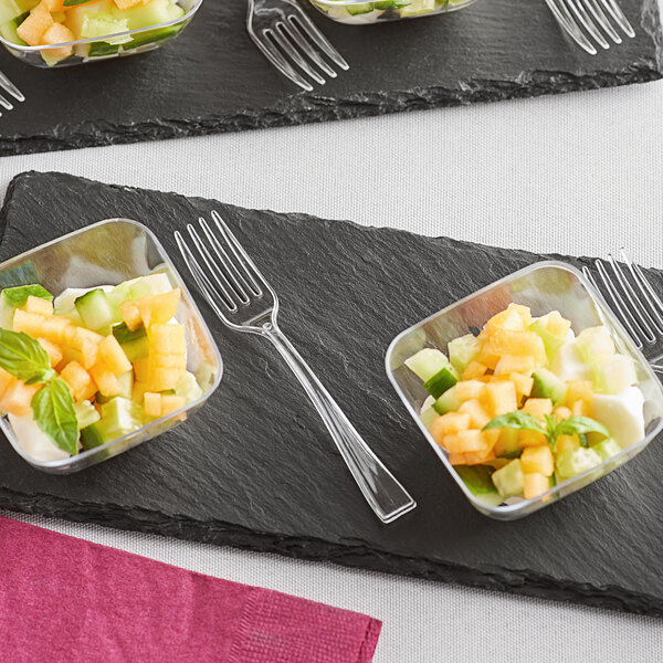 A Visions clear plastic tasting fork on a black surface with small bowls of fruit and vegetables.