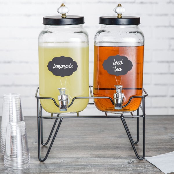 A Stylesetter glass beverage dispenser with metal stand and two dispensers filled with brown and yellow liquid.