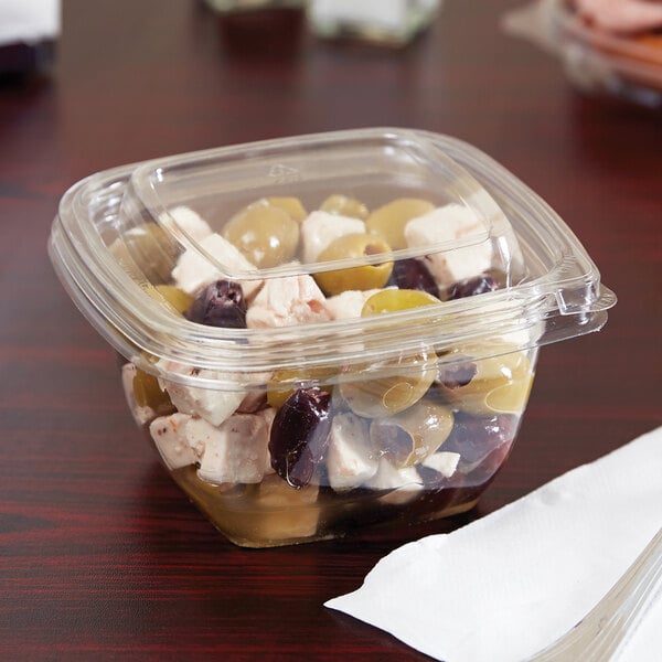 A plastic container with food in it on a table with olives and grapes.