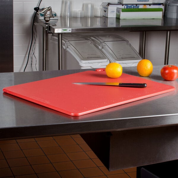 A San Jamar red cutting board on a counter with oranges and a knife.