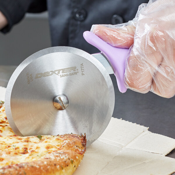 A person using a Dexter-Russell purple handled pizza cutter to cut a slice of pizza.