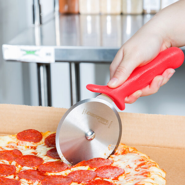 A hand with a red Dexter-Russell pizza cutter cutting a pizza.