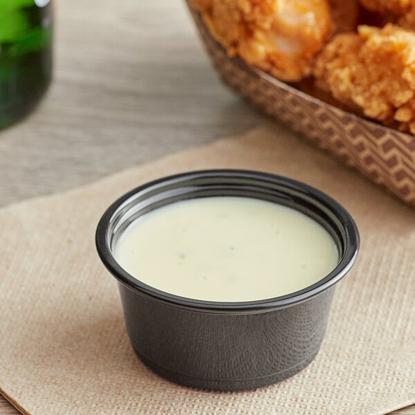 A bowl of white sauce next to fried chicken wings in a Choice black plastic souffle cup.
