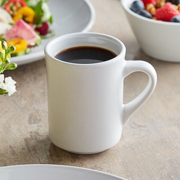 An Acopa bright white stoneware mug with a drink in it next to a bowl of fruit.