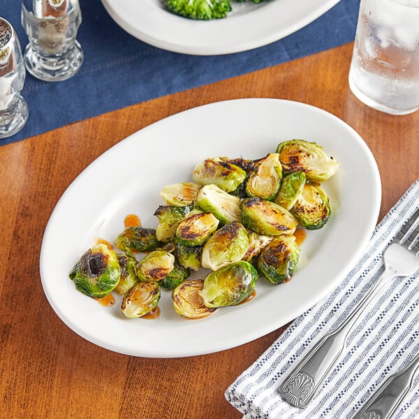 A table set with a plate of roasted brussels sprouts on an Acopa bright white rolled edge oval platter with a fork and a glass of ice.